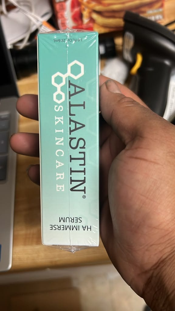 A hand holding a mint green box of ALASTIN SKINCARE HA IMMERSER Serum, with a cluttered desk background.